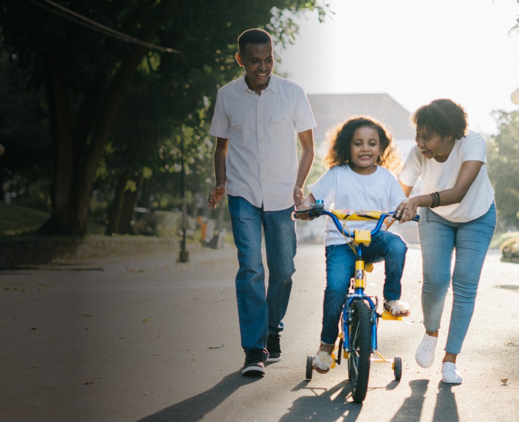 Two primary caregivers excitedly teaching a child how to ride a bike