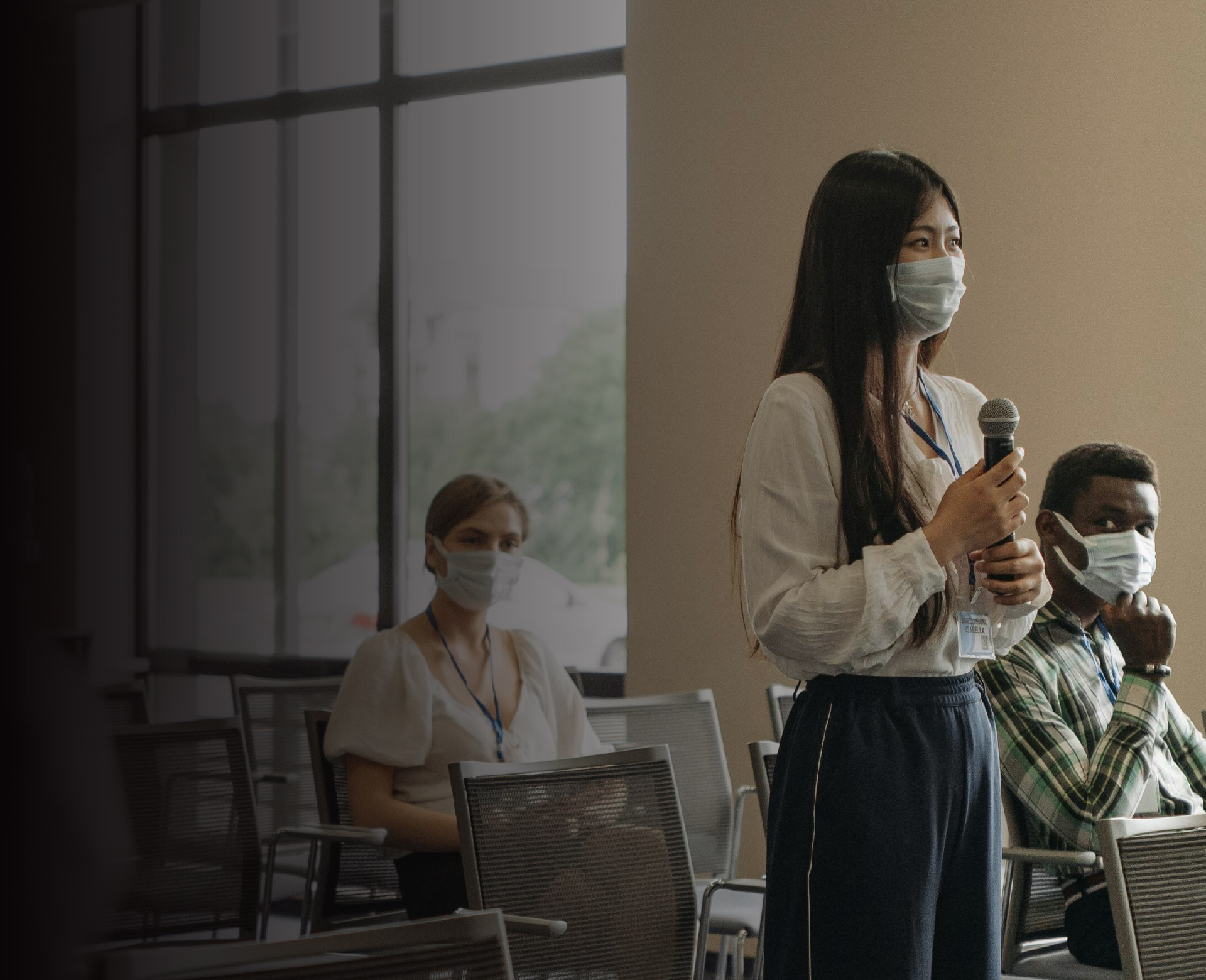 woman wearing a mask, standing up and giving a speech while holding a microphone in her hand